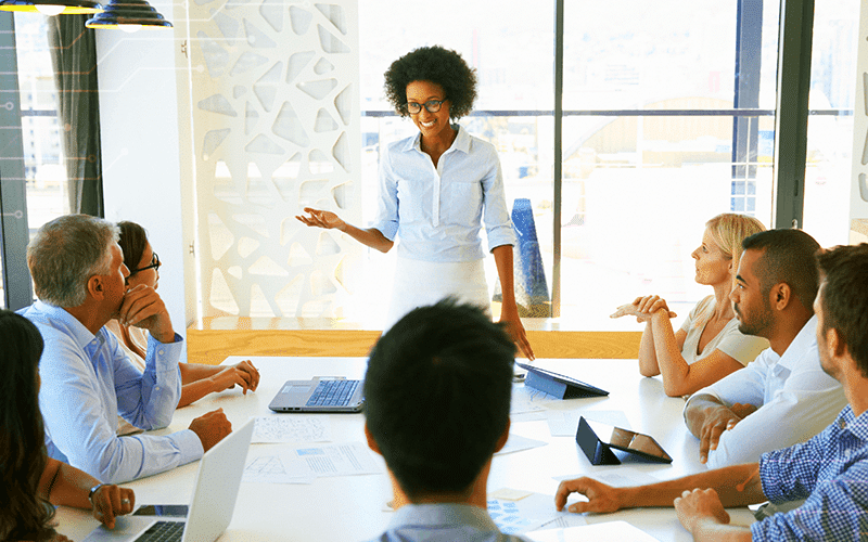Professional woman speaking to table of individuals at a meeting.